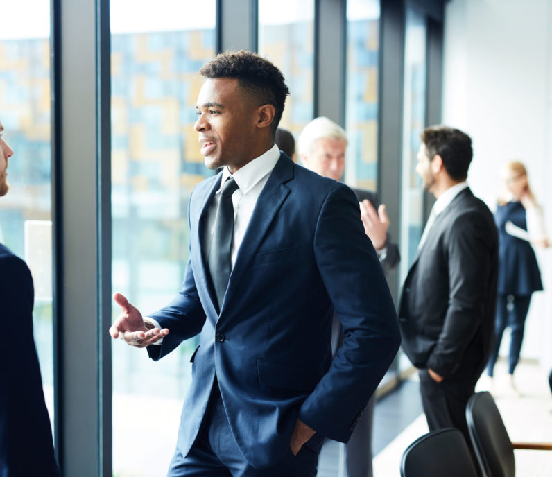 Two intercultural delegates and their colleagues on background discussing possible questions for speakers before political summit in conference hall