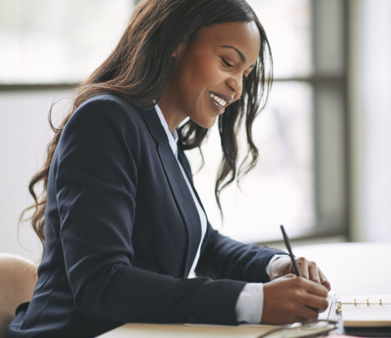 Smiling young African American businesswoman writing down notes in her day planner while sitting at a table in an office