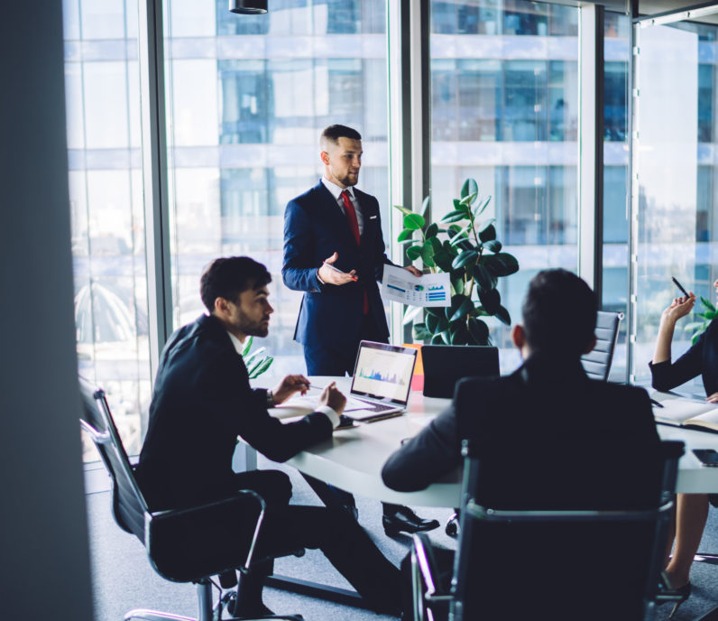 Confident successful businessman in formal black suit standing next to colleagues and analyzing financial report during conference in modern workspace